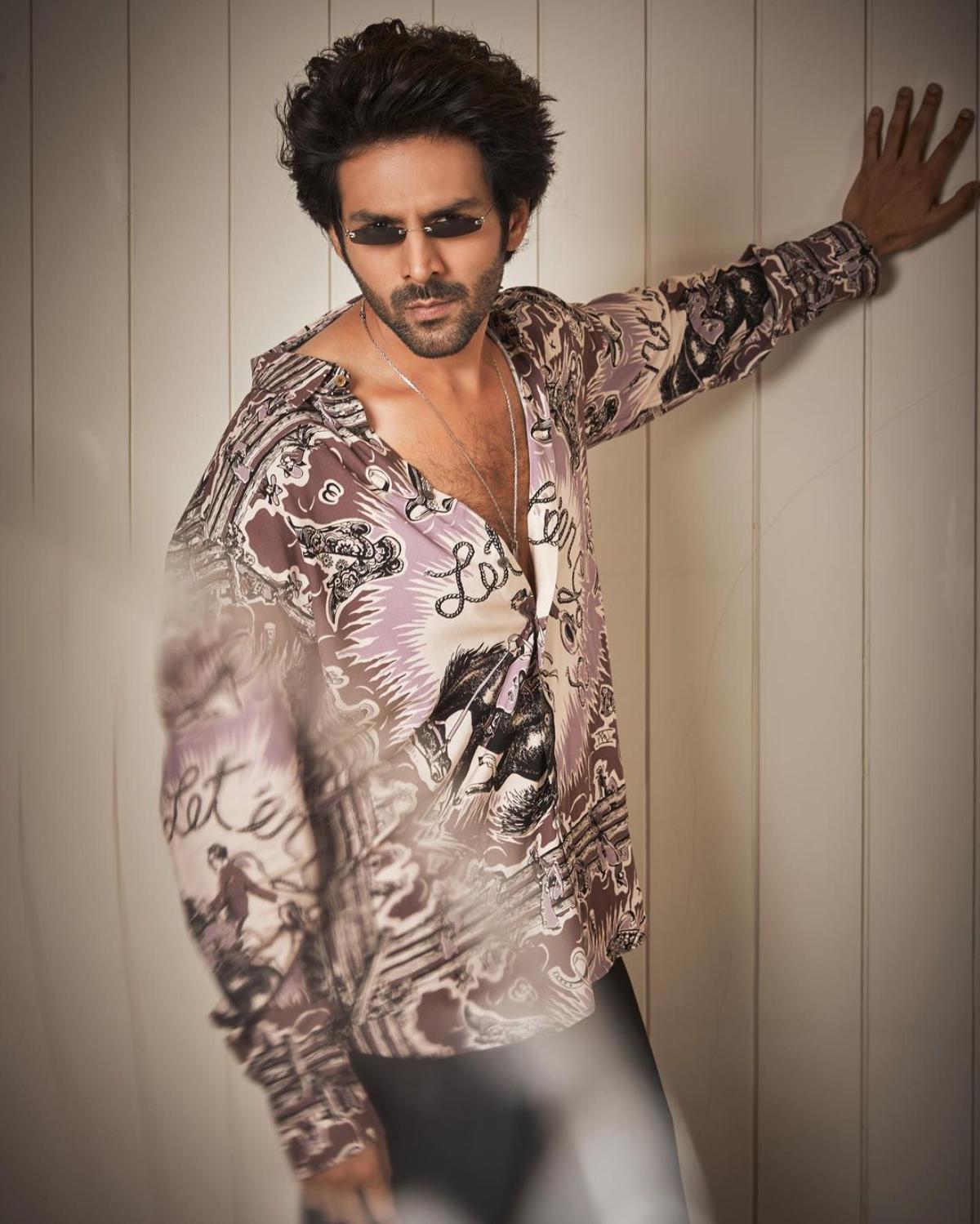 A printed shirt? Yes, please!
Well, well, well... men, if there's one ultimate party look of Kartik Aaryan from where you can take your fashion inspo from, it is THIS! A light coffee brown printed shirt with purple, white and black colour splattered all over, Kartik looks like a true-blue party animal who owns the dance floor with his killer moves. The actor who paired his printed shirt with something which appears to be a black skinny jeans, raised the temperature as he left the 4-5 buttons of his shirt undone. To take his hotness a notch higher, the actor wore his favourite, black sleek glasses. For the accessories, Kartik wore nothing but a long silver chain with a locket. With a sexy printed shirt like Kartik, we are sure you boys will make heads turn at parties.  